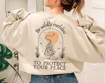 Può includere: Una felpa color crema con una grafica di una tigre con il testo "Be wildly ruthless to protect your peace" stampato sul retro.