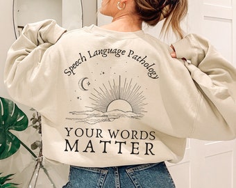 Puede incluir: Una sudadera beige con el texto "Speech Language Pathology" y un gráfico de sol y luna. El texto "Your Words Matter" está impreso debajo del gráfico.