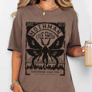 Pode incluir: Camiseta marrom com um gr&aacute;fico preto de Mothman, o texto "Mothman Live Show" e "Screaming Since 1966". O design inclui uma ponte e uma silhueta de floresta. A camiseta &eacute; de estilo casual e confort&aacute;vel.