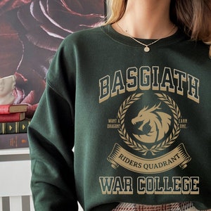 Op de afbeelding: Een donkergroen sweatshirt met een gouden grafische print die "Basgiath Riders Quadrant War College" zegt. De afbeelding toont een draak en een lauwerkrans.