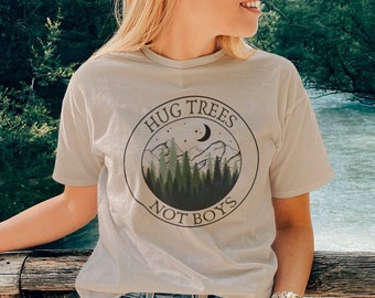Puede incluir: Una camiseta beige claro con un estampado gráfico de un círculo con el texto "Hug Trees Not Boys" y una escena de montaña con árboles y una luna creciente.
