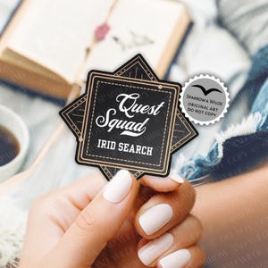 May include: A black and gold sticker with the words "Quest Squad" and "IRID SEARCH" in white. The sticker is held in a hand with white nail polish, with a book and a cup of coffee in the background.