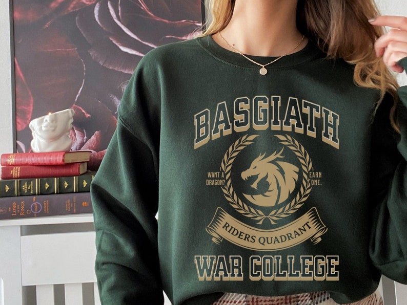 Op de afbeelding: Een donkergroen sweatshirt met een gouden grafische print die "Basgiath Riders Quadrant War College" zegt. De afbeelding toont een draak en een lauwerkrans.