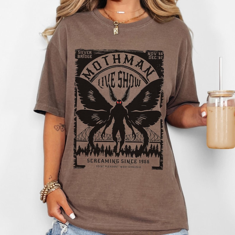 Pode incluir: Camiseta marrom com um gr&aacute;fico preto de Mothman, o texto "Mothman Live Show" e "Screaming Since 1966". O design inclui uma ponte e uma silhueta de floresta. A camiseta &eacute; de estilo casual e confort&aacute;vel.