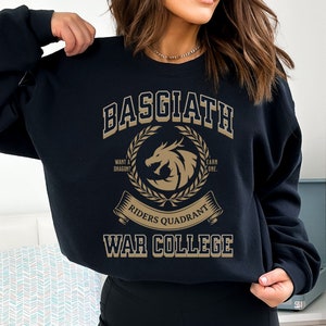 Op de afbeelding: Zwart sweatshirt met een gouden grafisch ontwerp met de tekst "Basgiath Riders Quadrant War College". De grafiek toont een draak met een krans eromheen en de tekst "Wil je een draak? Verdien er een."