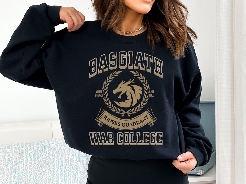 Op de afbeelding: Zwart sweatshirt met een gouden grafisch ontwerp met de tekst "Basgiath Riders Quadrant War College". De grafiek toont een draak met een krans eromheen en de tekst "Wil je een draak? Verdien er een."