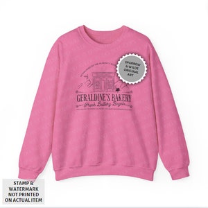 Puede incluir: Sudadera rosa con un dise&ntilde;o gr&aacute;fico negro de una panader&iacute;a llamada "Geraldine's Bakery" con el texto "Fresh Buttery Bagels". El gr&aacute;fico tambi&eacute;n incluye el texto "Established by the Almighty Sovereign" y "A Mile-try to Zoning Academy. How About Our Generous Discount?"