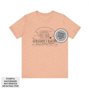 May include: A peach-coloured t-shirt featuring a black and white illustration of a bakery called "Geraldine's Bakery: Fresh Buttery Bagels". The illustration includes a shop front with a sign that reads "Geraldine's Bakery", a mountain range, and two birds. The text "Established by the Almonty Sovereign Society" appears above the illustration. The text "Free Delivery to Edgar Academy - Ask About Supernumerary Discount" appears below the illustration. A grey circle with the text "Sparrow & Wilde Original Art" appears in the top right corner of the image.