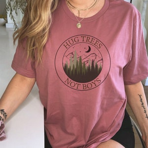 May include: A pink t-shirt with a graphic of a mountain range, trees, a crescent moon, and stars. The graphic is in a circle with the text "Hug Trees Not Boys".