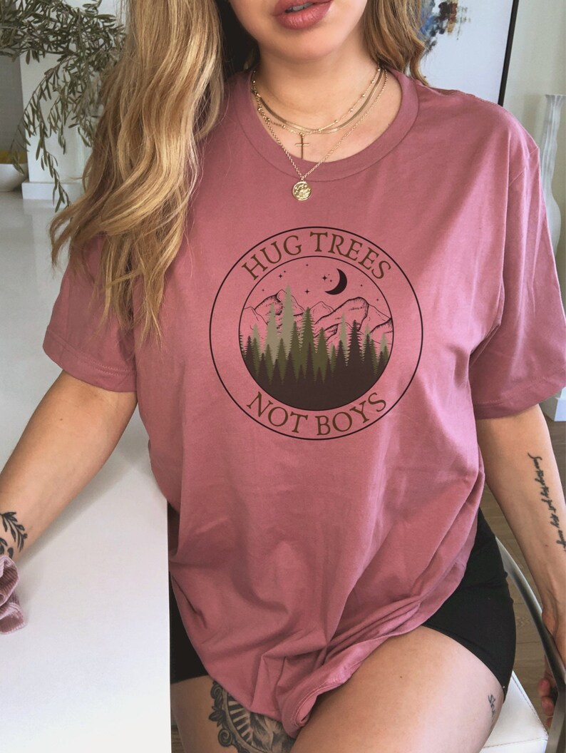 May include: A pink t-shirt with a graphic of a mountain range, trees, a crescent moon, and stars. The graphic is in a circle with the text "Hug Trees Not Boys".