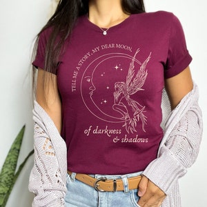 Op de afbeelding: Een maroon t-shirt met een wit grafisch ontwerp van een fee die op een halve maan zit met de tekst "Tell me a story, my dear moon, of darkness & shadows".