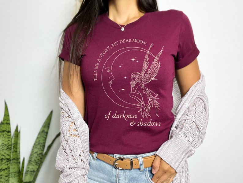 Op de afbeelding: Een maroon t-shirt met een wit grafisch ontwerp van een fee die op een halve maan zit met de tekst "Tell me a story, my dear moon, of darkness & shadows".