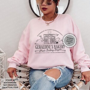 Puede incluir: Sudadera rosa con un dise&ntilde;o gr&aacute;fico negro de una panader&iacute;a llamada "Geraldine's Bakery" con el texto "Fresh Buttery Bagels". El gr&aacute;fico tambi&eacute;n incluye un sol, p&aacute;jaros y monta&ntilde;as. El texto "Established by the Almighty Sovereign Society" est&aacute; por encima del gr&aacute;fico de la panader&iacute;a. El texto "Free delivery to Zodiac Academy - Ask about our Cerberus discount" est&aacute; debajo del gr&aacute;fico de la panader&iacute;a. Un c&iacute;rculo gris con el texto "Sparrow & Wilde Original Art" est&aacute; en la esquina superior derecha del gr&aacute;fico.