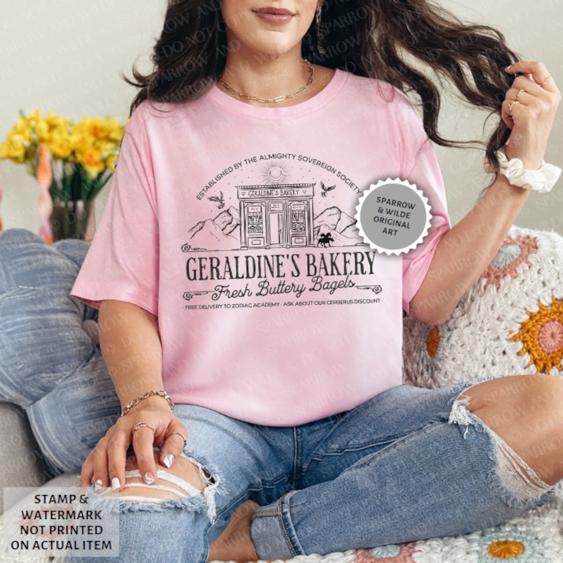 May include: A pink t-shirt featuring a black and white graphic design of a bakery called "Geraldine's Bakery" with the text "Fresh Buttery Bagels". The graphic also includes a small building with a sign that says "Geraldine's Bakery".