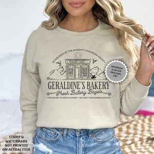 Puede incluir: Una sudadera beige con un dise&ntilde;o gr&aacute;fico negro de una panader&iacute;a llamada "Geraldine's Bakery" con el texto "Fresh Buttery Bagels". El gr&aacute;fico tambi&eacute;n incluye un sol, p&aacute;jaros, monta&ntilde;as y un caballo. El texto "Established by the Almighty Sovereign Society" est&aacute; por encima del gr&aacute;fico de la panader&iacute;a. El texto "Free delivery to Zodiac Academy. Ask about our Cerberus discount." est&aacute; debajo del gr&aacute;fico de la panader&iacute;a. El texto "Sparrow & Wilde Original Art" est&aacute; en un c&iacute;rculo en el lado derecho del gr&aacute;fico.