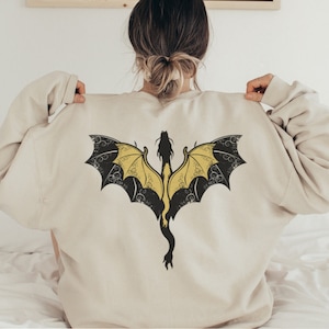 Puede incluir: Una sudadera de color crema con un dise&ntilde;o de drag&oacute;n negro y amarillo en la espalda.