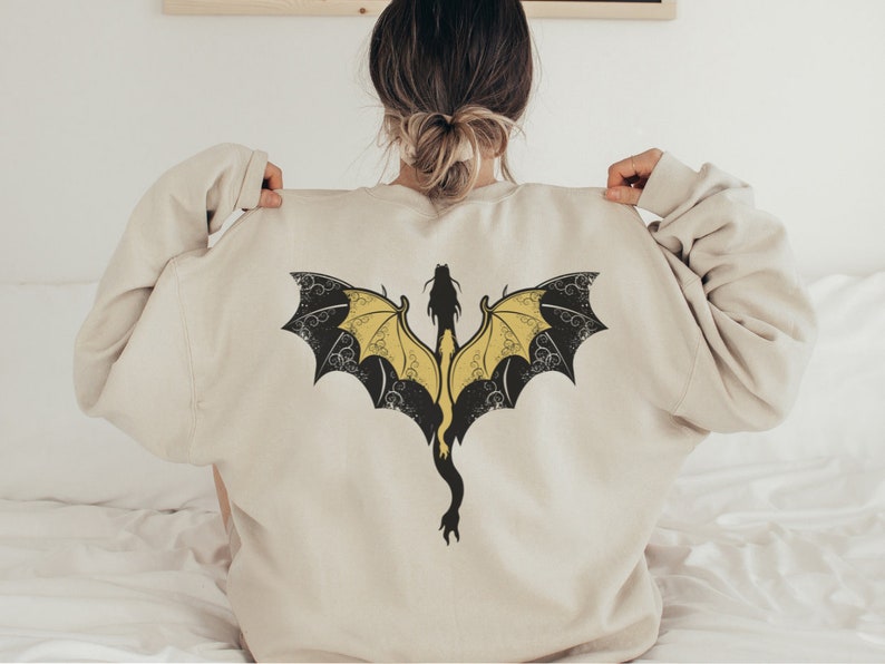 Puede incluir: Una sudadera de color crema con un dise&ntilde;o de drag&oacute;n negro y amarillo en la espalda.