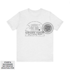 May include: A white t-shirt featuring a black and white illustration of a bakery called "Geraldine's Bakery" with the text "Fresh Buttery Bagels". The illustration includes a storefront with a sign that reads "Geraldine's Bakery". The text "Established by the Almighty Sovereign Society" is above the illustration. The text "Sparrow & Wilde Original Art" is in a circle with a starburst design.