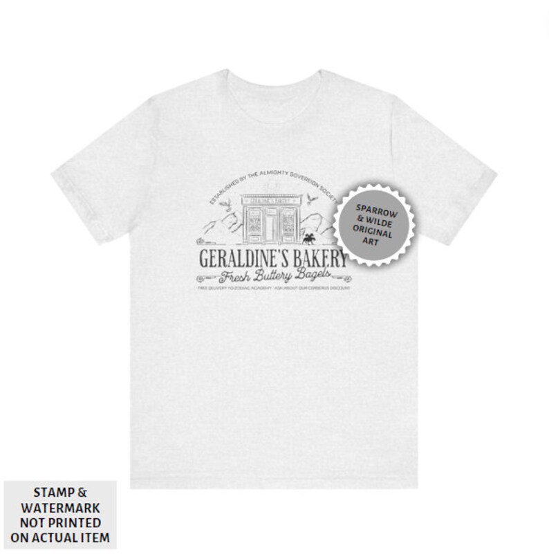 May include: A white t-shirt featuring a black and white illustration of a bakery called "Geraldine's Bakery" with the text "Fresh Buttery Bagels". The illustration includes a storefront with a sign that reads "Geraldine's Bakery". The text "Established by the Almighty Sovereign Society" is above the illustration. The text "Sparrow & Wilde Original Art" is in a circle with a starburst design.