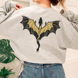 Puede incluir: Una sudadera gris con un dise&ntilde;o de drag&oacute;n negro y dorado en la espalda. El drag&oacute;n tiene las alas extendidas y una cola larga.