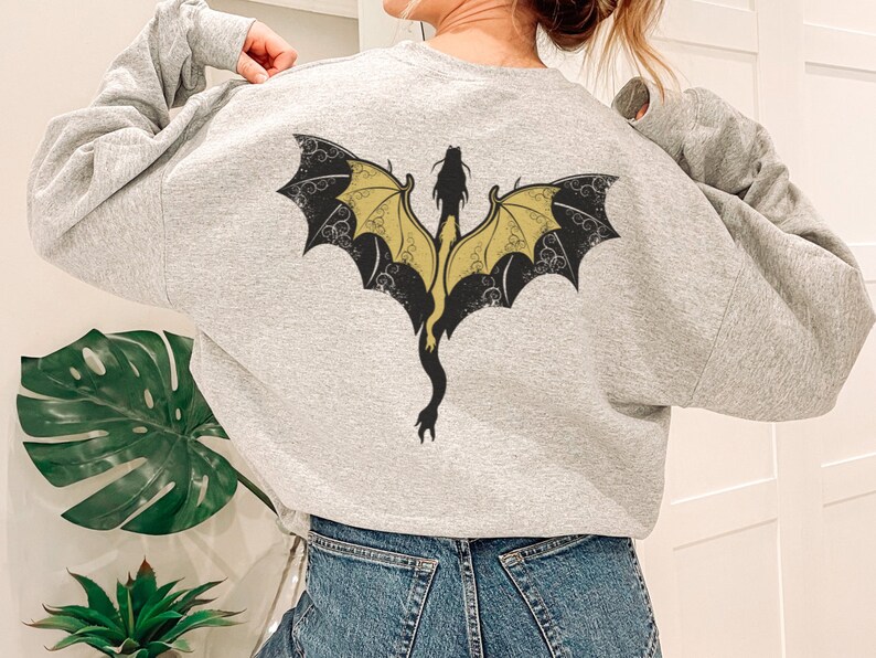 Puede incluir: Una sudadera gris con un dise&ntilde;o de drag&oacute;n negro y dorado en la espalda. El drag&oacute;n tiene las alas extendidas y una cola larga.