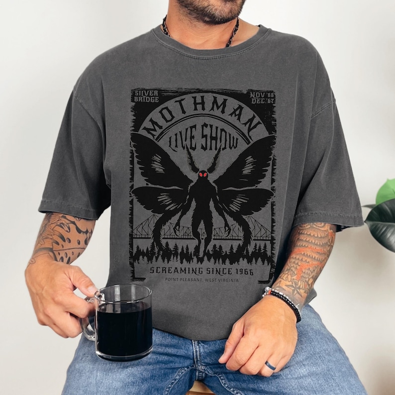 Pode incluir: Camiseta cinza escuro com um gr&aacute;fico Mothman Live Show. O design inclui uma silhueta de Mothman com grandes asas, a Silver Bridge e o texto "Screaming Since 1966". A camiseta &eacute; usada com jeans azul.