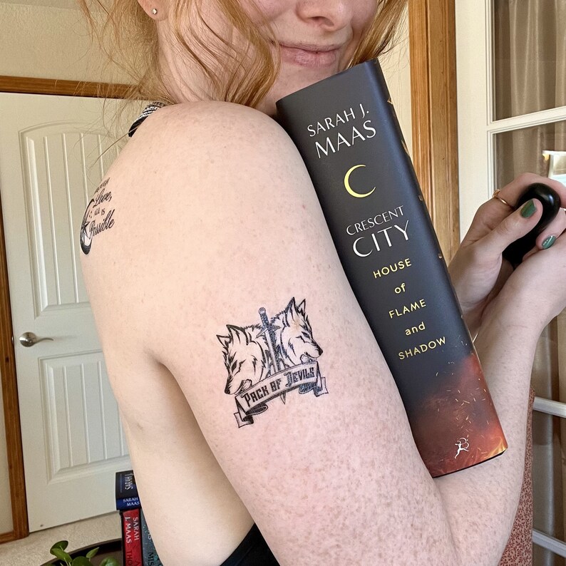 May include: A person's arm with a temporary tattoo of two wolves with a sword and the text "Pack of Devils". The person is holding a book titled "Crescent City, House of Flame and Shadow" by Sarah J. Maas.