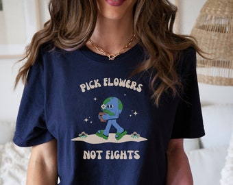 Puede incluir: Una camiseta azul marino con un gráfico de una Tierra de dibujos animados sosteniendo una maceta. El texto "PICK FLOWERS NOT FIGHTS" está impreso en la camiseta.