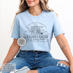 May include: Light blue t-shirt featuring a graphic of a bakery storefront with the text "Geraldine's Bakery Fresh Buttery Bagels" and the text "Established by the Almighty Sovereign Society" above the storefront. The graphic also includes mountains, birds, and a sun.