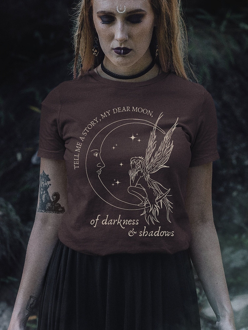 Op de afbeelding: Een bruin t-shirt met een afbeelding van een halve maan en een fee met de tekst "Tell me a story, my dear moon, of darkness & shadows".