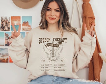 Puede incluir: Una sudadera beige con un gráfico marrón de una guitarra con alas y el texto "Speech Therapy World Tour". El gráfico también incluye una lista de temas de terapia del habla, como la articulación, la fluidez, la voz, el lenguaje, la audición, la deglución, la cognición, lo social y las modalidades. El texto "Your Words Matter" también está incluido.