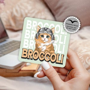 May include: Square sticker with a light green background featuring a cartoon cat wearing aviator goggles and headphones. The word "BROCCOLI" is printed in a retro font. The sticker also has the text "SPARROW & WILDE ORIGINAL ART DO NOT COPY".