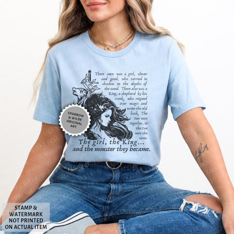 Puede incluir: Una camiseta azul claro con una ilustraci&oacute;n en blanco y negro de una ni&ntilde;a, un rey y un monstruo. El texto en la camiseta dice "There once was a girl, clever and good, who tarried in shadow in the depths of the wood. There also was a King, a shepherd by his crook, who reigned over magic and wrote the old book. The two were together, so the two were the same. The girl, the King... and the monster they became."