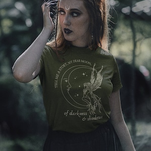 Op de afbeelding: Een groen T-shirt met een witte afbeelding van een halve maan met een fee die erop zit. De tekst op het shirt luidt "Tell me a story, my dear moon, of darkness & shadows".