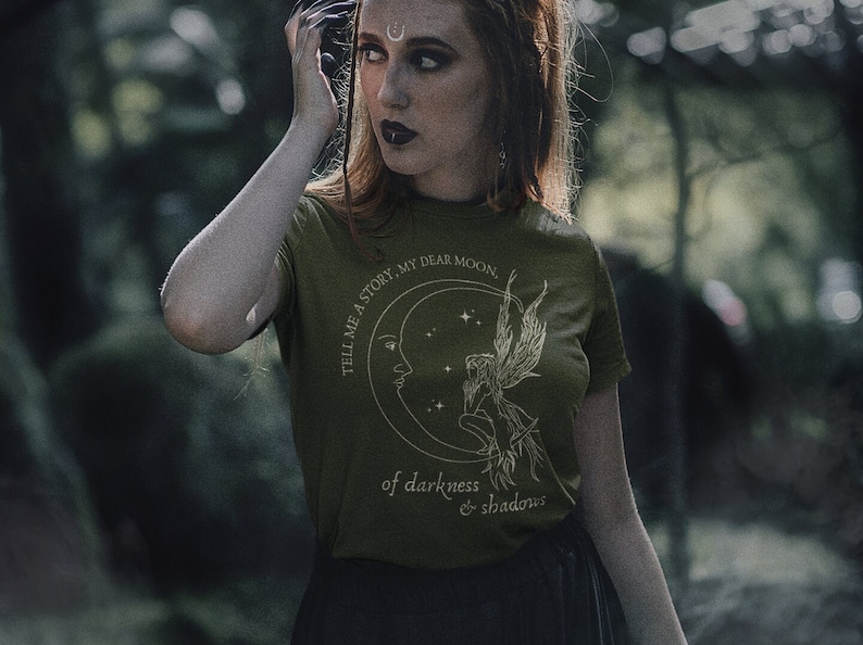 Op de afbeelding: Een groen T-shirt met een witte afbeelding van een halve maan met een fee die erop zit. De tekst op het shirt luidt "Tell me a story, my dear moon, of darkness & shadows".