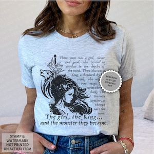Puede incluir: Una camiseta azul claro con una ilustraci&oacute;n en blanco y negro de una mujer con un lobo y una espada. El texto en la camiseta dice "There once was a girl, clever and good, who tarried in shadow in the depths of the wood. There also was a King, a shepherd by crook, who reigned over magic... The girl, the King... and the monster they became." El texto "Sparrow & Wilde Original Art" est&aacute; en un c&iacute;rculo en el lado derecho de la camiseta.