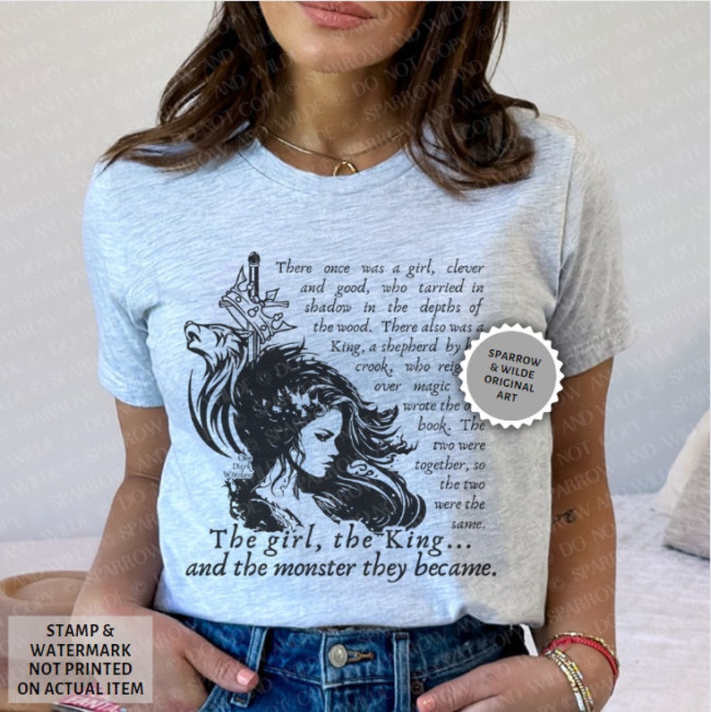 Puede incluir: Una camiseta azul claro con una ilustraci&oacute;n en blanco y negro de una mujer con un lobo y una espada. El texto en la camiseta dice "There once was a girl, clever and good, who tarried in shadow in the depths of the wood. There also was a King, a shepherd by crook, who reigned over magic... The girl, the King... and the monster they became." El texto "Sparrow & Wilde Original Art" est&aacute; en un c&iacute;rculo en el lado derecho de la camiseta.