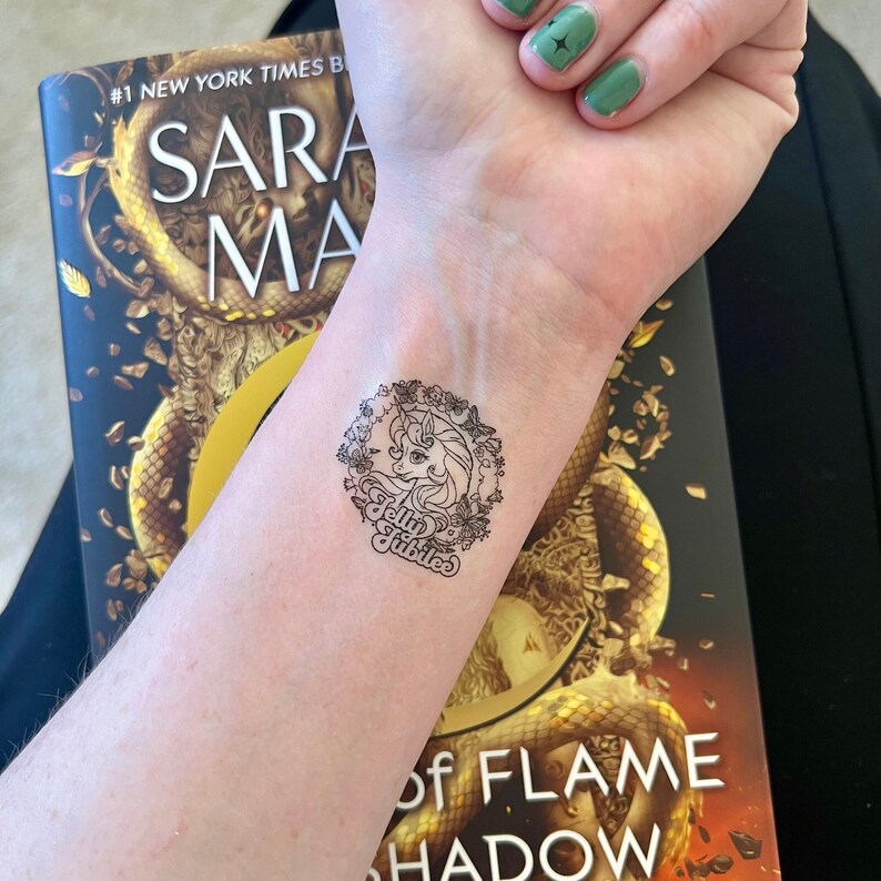 May include: A close-up of a person's wrist with a temporary tattoo of a unicorn with a floral wreath and the text "Jelly Jubilee". The tattoo is on the left wrist and the person is wearing green nail polish on their right hand.