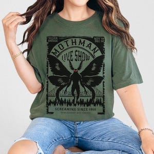Pode incluir: Camiseta verde-oliva com um gr&aacute;fico preto de Mothman, com o texto "Mothman Live Show" e "Screaming Since 1966". O design inclui uma ponte e uma silhueta de floresta. A camiseta tem um corte folgado.
