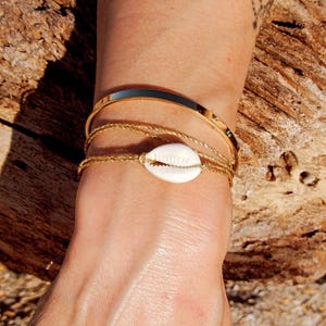 Peut inclure: Plusieurs bracelets dorés ornent un poignet. L'ensemble comprend un jonc doré épais et poli, une fine chaîne dorée et un bracelet chaîne doré orné d'un coquillage blanc. Les bijoux sont présentés sur un fond en bois naturel.