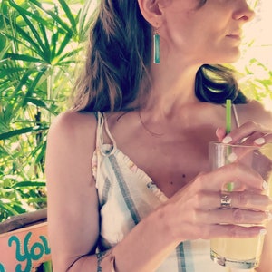Peut inclure: Une femme portant un haut rayé et un bracelet en or, avec de longues boucles d'oreilles turquoise, tient un verre de limonade avec une paille verte.