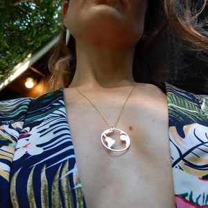 Puede incluir: Una mujer lleva una camisa de estampado floral azul y verde con un collar de cadena dorada con un colgante de plata en forma de mundo.