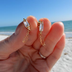 Peut inclure: Une paire de boucles d'oreilles dorées avec un cristal clair rectangulaire et une fine chaîne. Les boucles d'oreilles sont présentées sur une plage de sable blanc, d'eau bleue et d'un ciel clair.