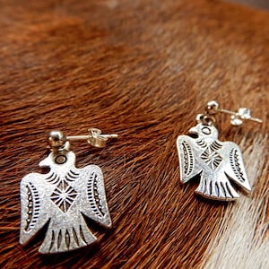 Peut inclure: Une paire de boucles d'oreilles argentées représentant un oiseau stylisé. Chaque boucle d'oreille présente une forme d'oiseau symétrique et détaillée avec des motifs gravés et une tige pour oreilles percées. Fond marron texturé.