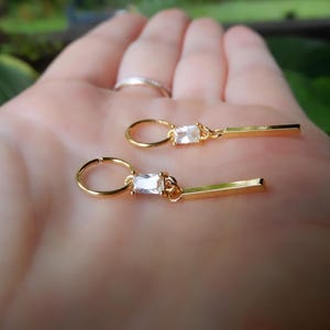 Peut inclure: Deux boucles d'oreilles dorées, chacune ornée d'une pierre précieuse rectangulaire et transparente et d'une petite barre dorée. Les boucles d'oreilles sont tenues dans une main, avec une bague visible. Design simple et élégant.