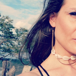 Peut inclure: Une femme aux longs cheveux noirs portant un collier ras du cou en coquillages blancs et des boucles d'oreilles en chaîne dorée. L'arrière-plan est une image floue d'une plage et du ciel.