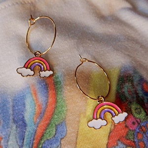 Peut inclure: Une paire de boucles d'oreilles créoles dorées avec des breloques arc-en-ciel. Chaque breloque représente un arc-en-ciel avec un schéma de couleurs rose, jaune et violet, et des nuages blancs de chaque côté.