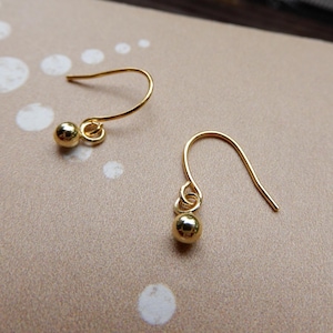 Peut inclure: Une paire de boucles d'oreilles dorées. Chaque boucle d'oreille est composée d'une petite perle ronde suspendue à un crochet incurvé. Les boucles d'oreilles sont présentées sur une surface marron clair avec des motifs circulaires blancs.