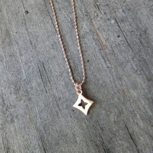Peut inclure: Un collier en or rose avec un petit pendentif en forme de losange ajouré. Le pendentif présente un motif à quatre pétales. La chaîne du collier est de style perlé fin. Le fond est une surface en bois vieilli.