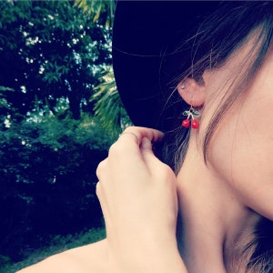 Peut inclure: Une femme portant un chapeau noir avec des boucles d'oreilles en forme de cerises. Les boucles d'oreilles sont en or avec des cerises rouges et des feuilles vertes.